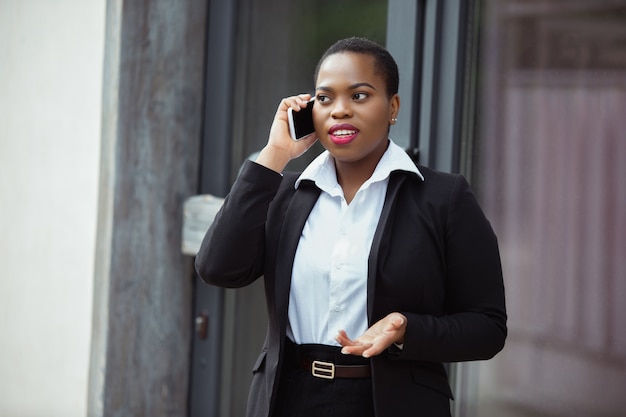 La donna d'affari afroamericana in abbigliamento da ufficio sorridente sembra sicura di parlare al telefono