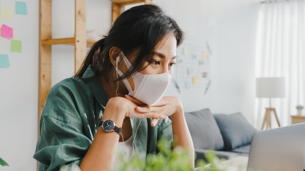 La donna d'affari asiatica che indossa una maschera medica utilizzando il laptop parla con i colleghi del piano in videochiamata mentre lavora da casa in soggiorno.