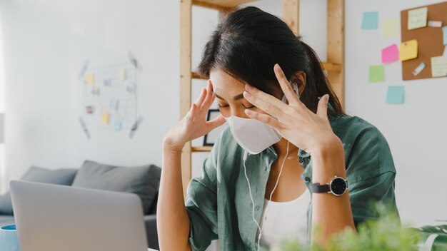 La donna d'affari asiatica che indossa una maschera medica utilizzando il laptop parla con i colleghi del piano in videochiamata mentre lavora da casa in soggiorno.