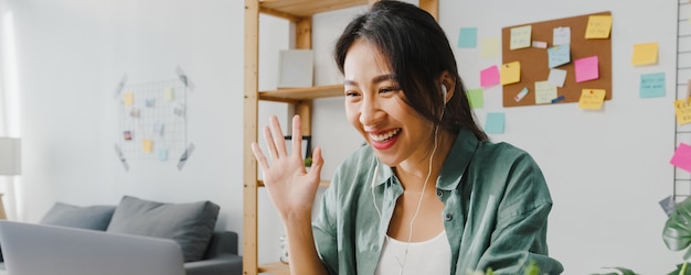 La donna d'affari asiatica che utilizza il laptop parla con i colleghi del piano in videochiamata mentre lavora in modo intelligente da casa in soggiorno