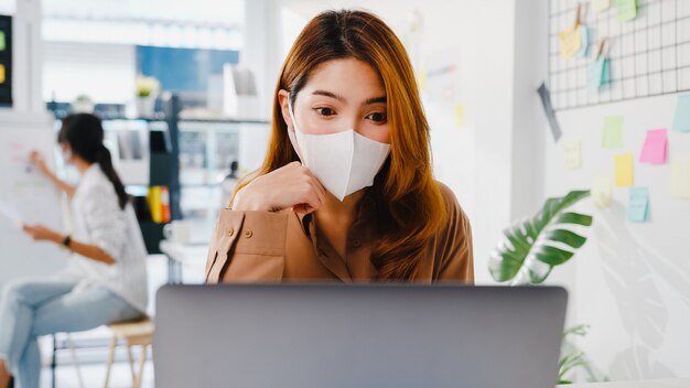 La donna d'affari asiatica indossa una maschera per il distanziamento sociale in una nuova situazione normale per la prevenzione dei virus durante l'utilizzo della presentazione del laptop ai colleghi sul piano in videochiamata mentre si lavora in ufficio.