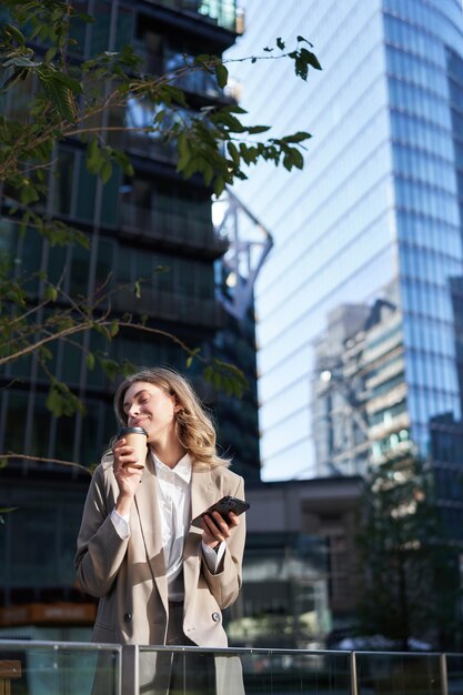 La donna d'affari entusiasta beve il suo caffè da asporto per strada tiene il telefono cellulare in piedi in beige