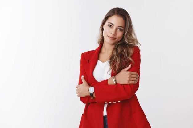 La donna d'affari può sembrare tenera. Studio shot attraente femminile giovane donna lavoratrice che indossa una giacca rossa che si abbraccia sorridente carino testa inclinabile guardando morbido, dipendente di sesso femminile che frequenta l'ufficio-festa