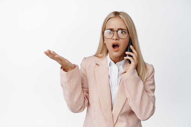 La donna d'affari scioccata riceve cattive notizie sul telefono che scrolla le spalle e sembra frustrata in piedi in tuta su sfondo bianco