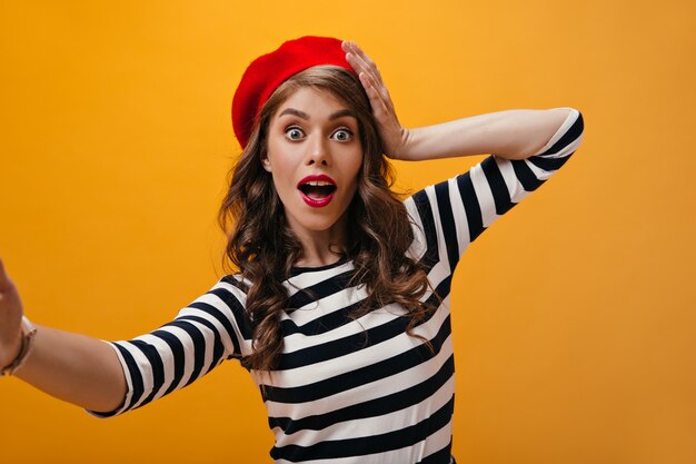 La donna dagli occhi aperti in berretto rosso fa selfie. Ragazza riccia sorpresa con cappello luminoso in maglioni moderni a strisce che esamina la macchina fotografica.