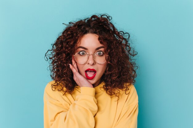 La donna dagli occhi azzurri con le labbra rosse guarda la telecamera con la bocca aperta per la sorpresa. Ragazza riccia in maglione giallo in posa sullo spazio blu.