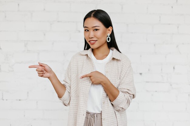 La donna dagli occhi marroni in elegante cardigan abs pantaloni beige sorride, esamina la macchina fotografica e indica il posto per il testo sul muro di mattoni bianchi