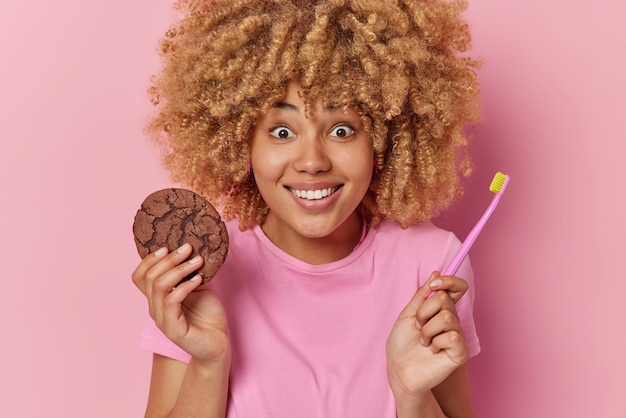 La donna dai capelli ricci allegra e sorpresa tiene un delizioso biscotto al cioccolato mangia cibo dannoso con una cattiva influenza sui denti pone con lo spazzolino da denti indossa una maglietta casual isolata su sfondo rosa