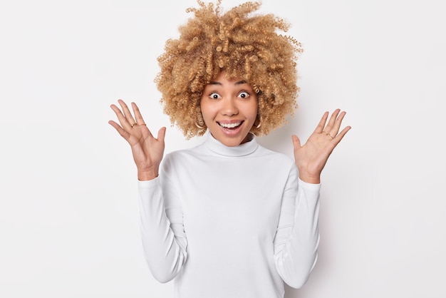 La donna dai capelli ricci eccitata gioiosa tiene i palmi sollevati esclama dalla felicità reagisce su qualcosa di fantastico indossa il dolcevita isolato su sfondo bianco Emozioni umane e concetto di reazioni