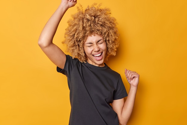 La donna dai capelli ricci emotiva stringe i pugni in aria si sente eccitata esclama di gioia indossa una maglietta nera casual isolata su sfondo giallo celebra il successo o il successo Sì, l'ho fatto finalmente