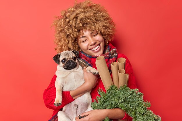 La donna dai capelli ricci felice ha relazioni amichevoli con il cane carlino preferito tiene una corona verde e la carta arrotolata indossa un maglione e una sciarpa intorno al collo isolati su sfondo rosso. Capodanno.