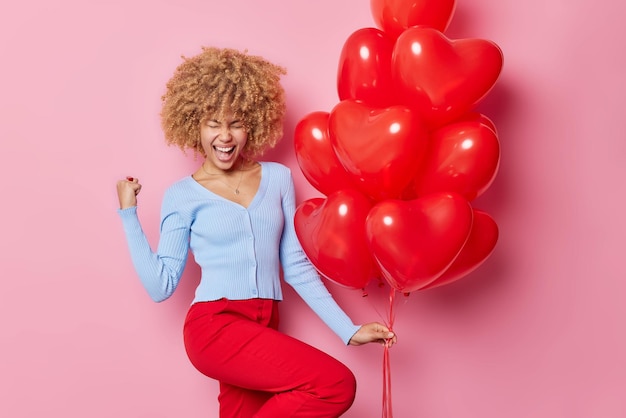 La donna dai capelli ricci felicissima esclama forte stringe il pugno celebra l'occasione speciale indossa un maglione casual e pantaloni rossi tiene un mazzo di palloncini a cuore posa su sfondo rosa Buone vacanze