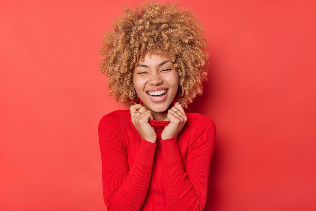 La donna dai capelli ricci gioiosa felice sorride ampiamente mostra i denti bianchi esprime emozioni sincere autentiche tiene le mani sul colletto del maglione isolato su sfondo rosso. Concetto di gioia e divertimento della gente