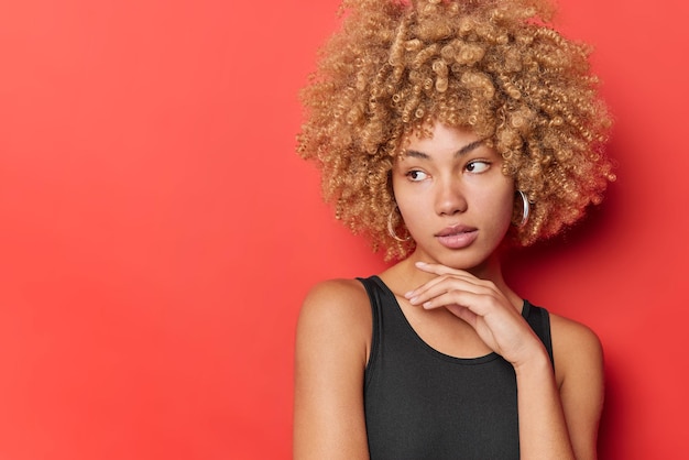 La donna dai capelli ricci pensierosa con la bellezza naturale tocca la mascella concentrata da parte vestita con una maglietta nera immersa nei pensieri isolati su sfondo rosso copia spazio per i tuoi contenuti promozionali.