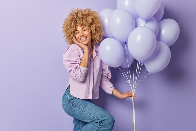 La donna dai capelli ricci positiva celebra l'occasione speciale si sente molto felice riceve congratulazioni tiene un mazzo di palloncini gonfiati indossa camicia e jeans isolati su sfondo viola Tempo di festa