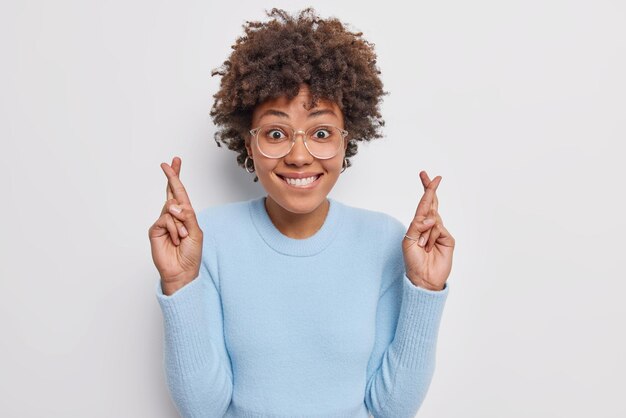 La donna dai capelli ricci positiva speranzosa prega e desidera desidera sorride felicemente indossa occhiali casual maglione blu ha grandi speranze attende risultati pose su sfondo bianco. Linguaggio del corpo