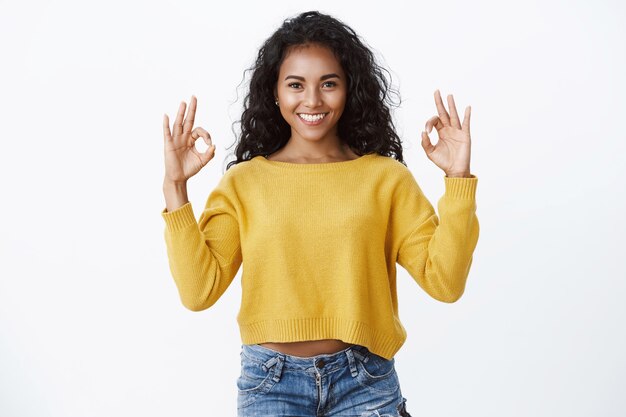 La donna dai capelli ricci sicura di sé in maglione giallo dà un feedback positivo, mostra ok, buono, gesto eccellente sorridendo con approvazione, sembrando soddisfatto