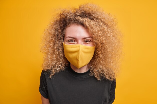 La donna dai capelli ricci soddisfatta indossa una maschera protettiva per prevenire la diffusione del coronavirus vestita con una maglietta nera casual esprime emozioni positive isolate sul muro giallo. Tempo di quarantena