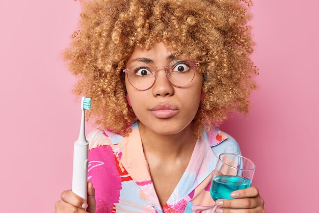 La donna dai capelli ricci sorpresa che va a lavarsi i denti tiene uno spazzolino elettrico e un bicchiere di collutorio indossa occhiali per una buona vista indossa una camicia casual isolata su sfondo rosa Igiene quotidiana