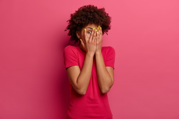 La donna dai capelli ricci spaventata copre il viso con i palmi delle mani, si arrabbia di qualcosa, esprime paura, guarda e sbircia tra le dita, si nasconde, indossa una maglietta casual, posa contro un muro roseo