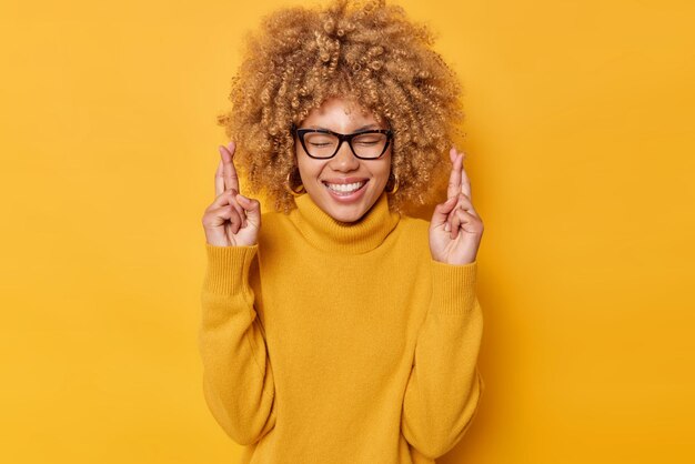 La donna dai capelli ricci superstiziosa tiene le dita incrociate per buona fortuna sorride positivamente tiene gli occhi chiusi indossa occhiali e maglione casual isolato su sfondo giallo. Concetto di linguaggio del corpo