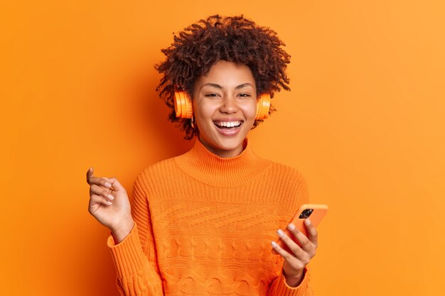 La donna dai capelli riccia allegra tiene lo smartphone moderno alza i sorrisi della mano ampiamente