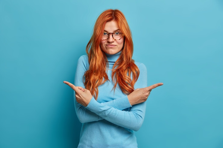 Foto gratuito la donna dai capelli rossi dubbiosa sceglie tra due modi in cui incrocia le braccia sul corpo indica su lati diversi vestiti con abiti casual.