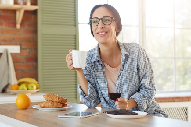 La donna dall'aspetto piacevole indossa una camicia casual e occhiali alla moda, tiene la tazza con il caffè e mangia cioccolato dolce,