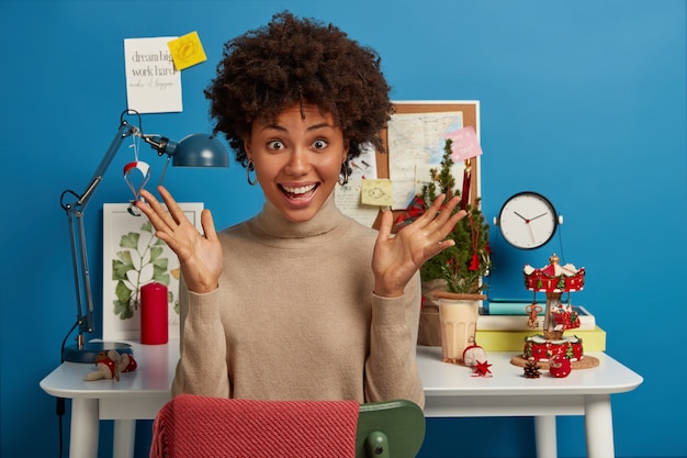 La donna dalla pelle scura allegra e allegra ha entrambi i palmi sollevati, si siede al desktop con l'albero di Natale e altri attributi delle vacanze