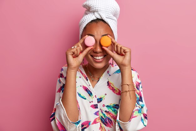 La donna dalla pelle scura sorridente positiva copre gli occhi con deliziosi amaretti dolci, ottiene calorie, si diverte a mangiare cibi dolci, indossa un asciugamano da bagno sulla testa, vestaglia domestica casual. Donne e concetto di dieta