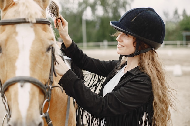 La donna del cavaliere pettina il suo cavallo in un ranch. La donna ha i capelli lunghi e vestiti neri. Equestre femminile che tocca il suo cavallo marrone.