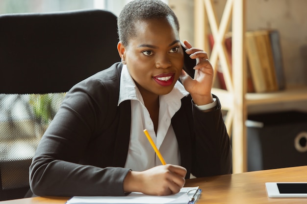 La donna di affari afroamericana in abbigliamento dell'ufficio che sorride, sembra sicura e felice, occupata