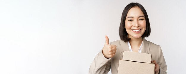 La donna di affari asiatica sorridente che mostra i pollici in su e le scatole con le merci di consegna prepara l'ordine per il cliente che si leva in piedi sopra fondo bianco