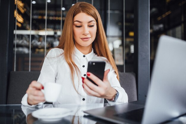 La donna di affari vestita in camicetta bianca e con capelli lisci lunghi si siede al tavolo con una tazza di caffè e guarda nel telefono con sguardo compiaciuto