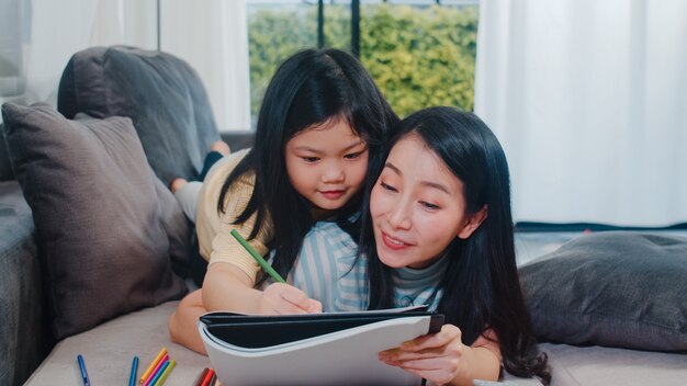 La donna di mezza età asiatica insegna a sua figlia a fare i compiti e disegnare a casa. Stile di vita madre e bambino divertimento felice trascorrere del tempo insieme nel salotto di casa moderna la sera.