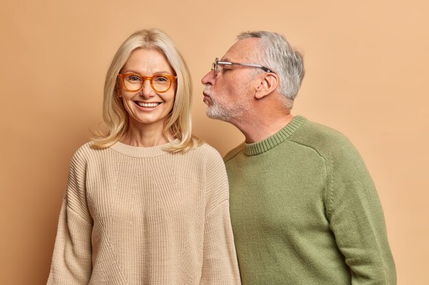 La donna di mezza età europea allegra sorride delicatamente mentre riceve il bacio dal marito ha buoni rapporti si amano per lungo tempo isolati sopra il muro marrone