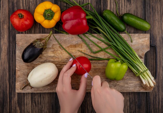 La donna di vista superiore taglia il pomodoro con le melanzane bianche e nere delle cipolle verdi su un tagliere con i peperoni colorati su un fondo di legno