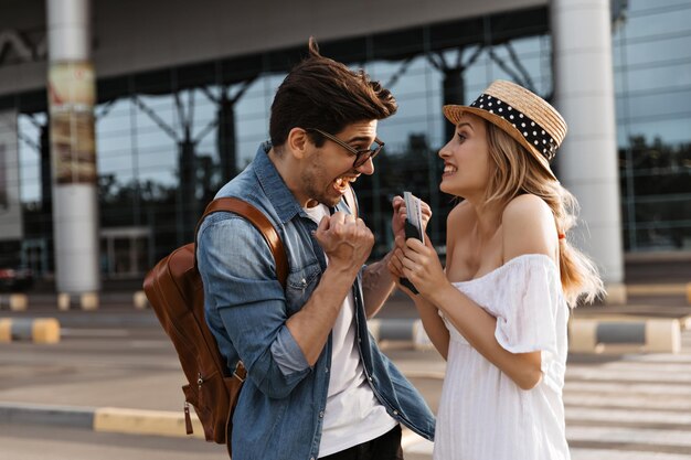 La donna e l'uomo biondi felici super eccitati si rallegrano vicino all'aeroporto I turisti non vedono l'ora di un nuovo viaggio Ragazza con cappello e ragazzo con occhiali da sole tengono passaporto e biglietti