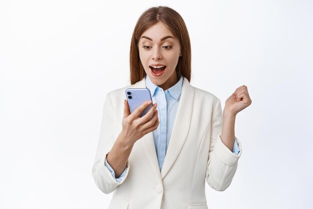 La donna eccitata dell'amministratore delegato di successo guarda al telefono leggere le notizie vincendo soldi online trionfando dal successo nell'applicazione sfondo bianco