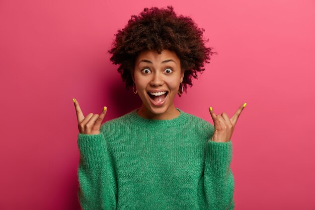 La donna eccitata riccia eccitata positiva fa un segno rock, trascorre il tempo libero al concerto della band preferita, esclama con gioia mentre ascolta una canzone fantastica, vestita con un maglione verde, posa su un muro rosa brillante