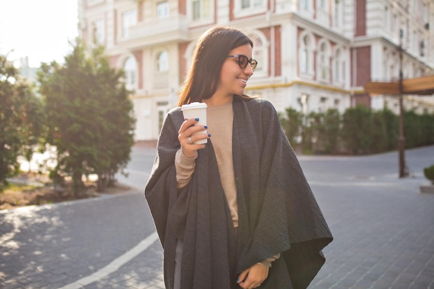 La donna elegante della stampa in poncho grigio e occhiali sta bevendo caffè e guardando da parte sulla strada soleggiata