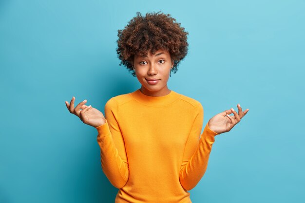 La donna esitante ha un'espressione perplessa alza le spalle e sembra confusa vestita in abiti casual isolati sopra il muro blu