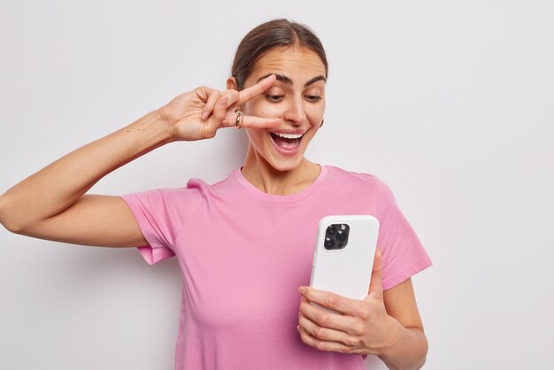 La donna europea bruna positiva fa un gesto di pace sull'occhio tiene il telefono cellulare e fa abiti da selfie in una maglietta rosa casual pone su sfondo bianco Concetto di divertimento e tecnologia della gente