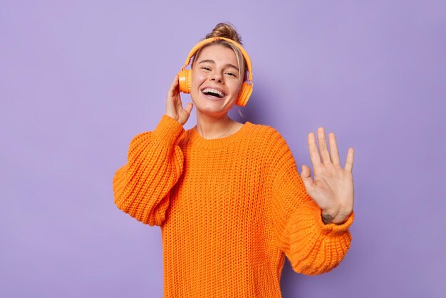 La donna europea spensierata positiva balla con il ritmo della musica ascolta la canzone preferita tramite le cuffie indossa un maglione arancione lavorato a maglia sorrisi ampiamente isolati su sfondo viola sciocca in giro