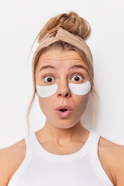 La donna europea stupita e stupita applica una maschera per gli occhi idratante antietà si sottopone a un trattamento per la pelle indossa la fascia per la testa e le t-shirt casual con gli occhi buggati isolati su sfondo bianco.