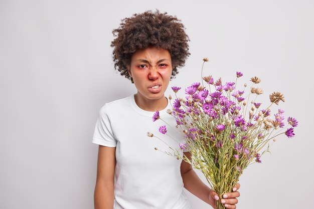 la donna fa un sorrisetto sul viso ha naso chiuso arrossamento intorno agli occhi reagisce al grilletto tiene il mazzo di fiori selvatici soffre di sintomi di febbre da fieno isolati su bianco