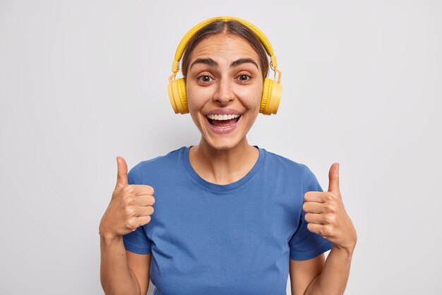 La donna felice approva qualcosa tiene i pollici in su consiglia di ascoltare questa traccia audio indossa le cuffie stereo gialle sulle orecchie vestite con una maglietta blu isolata su sfondo bianco. Mi piace.