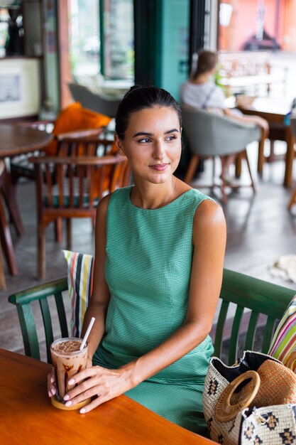 La donna felice calma elegante sveglia in vestito verde da estate si siede con il caffè nella caffetteria godendo la mattina