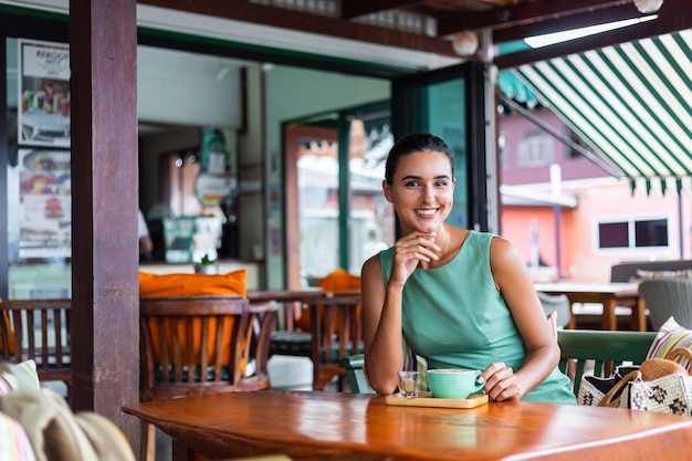 La donna felice calma elegante sveglia in vestito verde da estate si siede con il caffè nella caffetteria godendo la mattina