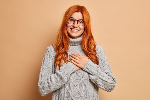 La donna felice della testarossa tiene le mani al petto fa sorrisi di gesto di gratitudine indossa felicemente il maglione grigio lavorato a maglia.